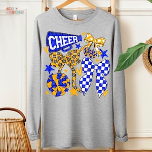 May include: A long-sleeved gray sweatshirt featuring a cheerleading design. The design includes the word "CHEER" in blue, along with yellow and blue bows, stars, and pom-poms. The sweatshirt is hanging on a wooden hanger.