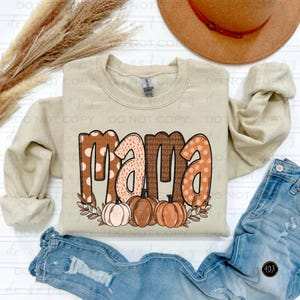 May include: A beige sweatshirt with the word "mama" in brown, orange, and white polka dot patterned letters. The letters are decorated with fall leaves and pumpkins.