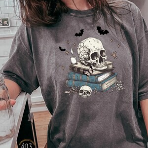 May include: A gray t-shirt with a graphic of a skull sitting on a stack of books. The skull has a candle on top of it and there are bats and stars around it.