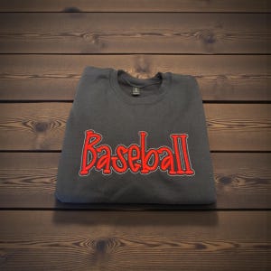 May include: A dark gray sweatshirt with the word "Baseball" embroidered in red with a white outline. The sweatshirt is folded and resting on a wooden surface.