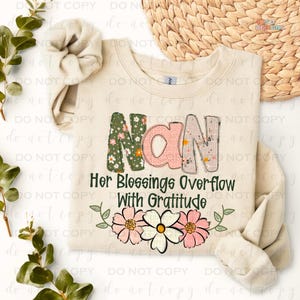 May include: A beige sweatshirt with the words "Nan Her Blessings Overflow With Gratitude" in green and pink letters with floral designs. The letters are surrounded by a floral border.