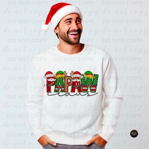 May include: White long-sleeved shirt with a Christmas design featuring the word "Papa" with elves and a reindeer. The word "Claus" is written below the word "Papa".