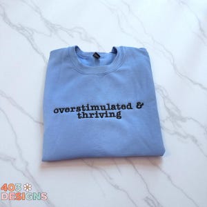 May include: A light blue sweatshirt with the words "overstimulated & thriving" embroidered in black. The sweatshirt is folded neatly on a white and gray marble surface. The brand logo "403 DESIGNS" is in the bottom left corner.