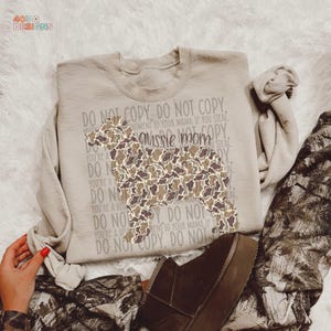 May include: A beige sweatshirt with a camouflage-patterned dog silhouette and the text "aussie mom." The words "DO NOT COPY" are printed around the dog. The sweatshirt is paired with camouflage pants and brown shoes.