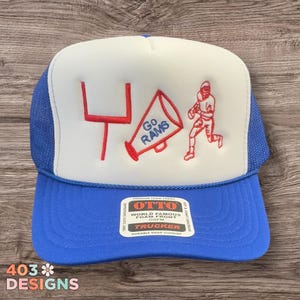 May include: A blue and white trucker hat with a football theme. The hat features a red goalpost, megaphone with "Go Rams" text, and a football player graphic. The hat has a snap closure and a "World Famous Foam Front" tag.
