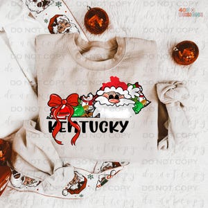 May include: A beige crewneck sweatshirt featuring a Christmas-themed design. The design includes a Santa Claus illustration, a red bow, and the word "Kentucky" in black. The design is in the shape of the state of Kentucky.