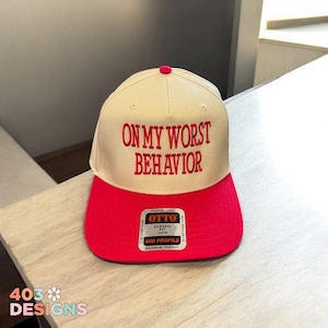 May include: A beige and red baseball cap with the text "ON MY WORST BEHAVIOR" embroidered in red on the front. The cap has a black label on the back that says "OTTO CLASSIC FIT OSFM MID PROFILE MAXIMUM COMFORT".