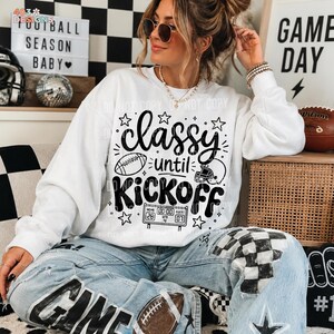 May include: A white long-sleeve sweatshirt featuring black graphic text that reads Classy until KICKOFF. The design includes illustrations of a football, a helmet, and a scoreboard displaying 00:00. The sweatshirt is worn with light blue jeans.