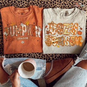 May include: Two fall-themed t-shirts. One is orange with "Pumpkin Season" text, the other is light gray with "Cozy Season" text. Both shirts have autumn-themed graphics. The image also shows a person holding a mug and wearing jeans and brown shoes.