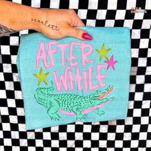 May include: A light blue t-shirt with the words "AFTER WHILE" in pink block letters, along with a green alligator illustration and yellow and pink star accents. The shirt is held up by a hand.