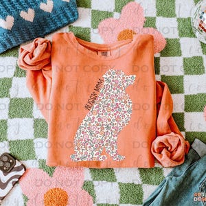 May include: A peach-colored sweatshirt with a floral patterned silhouette of an Australian Shepherd dog. The text "Aussie Mom" is printed on the dog's body.