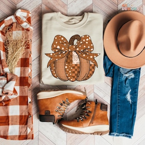 May include: A cream-colored sweatshirt with a pumpkin design, brown boots, a plaid shirt, a hat, and jeans. The pumpkin has a bow with polka dots and the text "TCO" repeated. The boots are brown with white soles.