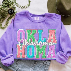 May include: Purple sweatshirt with a colorful glitter design that says "OKLAHOMA" in large letters.