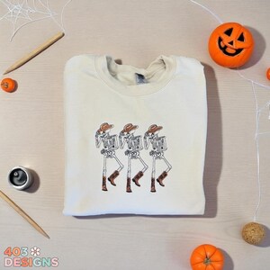 May include: Cream-colored sweatshirt featuring an embroidered design of three dancing skeletons wearing cowboy hats and boots. The design is in brown and black. The sweatshirt is on a light wood surface with Halloween decorations.