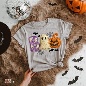 May include: A light gray t-shirt featuring a Halloween design. The design includes the word "BOO" in purple and yellow, a ghost, and a jack-o'-lantern. Black bats and spiderwebs are also included in the design. The shirt is tied at the waist.