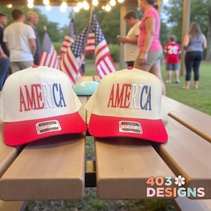 May include: Two white and red baseball caps with the word "AMERICA" embroidered on the front. The caps have a red brim and a white crown. The caps are sitting on a wooden table. American flags are in the background.