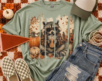 May include: A sage green t-shirt featuring a graphic of a raccoon wearing leopard print sunglasses and a plaid shirt while holding an iced coffee. The design includes autumn leaves, pumpkins, and a fence. The text 403 DESIGNS is in the corner.