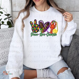 May include: White sweatshirt with a green, purple, and gold peace sign, heart, and crawfish design. The text "Peace Love Crawfish" is printed below the design.