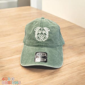 May include: A sage green dad hat with a white embroidered dog face design. The hat has a curved brim and an adjustable strap. The tag reads "OTTO Relaxed Fit Dad Hat".