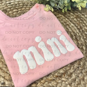 Op de afbeelding: Roze T-shirt met het woord "mini" in witte, opgevulde letters.