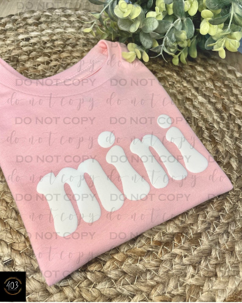 Op de afbeelding: Roze T-shirt met het woord "mini" in witte, opgevulde letters.