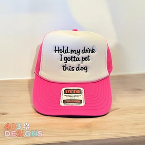 May include: Pink and white trucker hat with black embroidered text that reads "Hold my drink I gotta pet this dog". The hat has a black label with white text that reads "OTTO World Famous Foam Front OSFM Trucker Durable Snap Closure".
