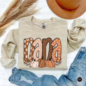 May include: A beige sweatshirt with the word "mama" printed in brown, orange, and white with a polka dot, leopard print, and textured pattern. The word is surrounded by a leafy garland with three pumpkins in orange, brown, and white.