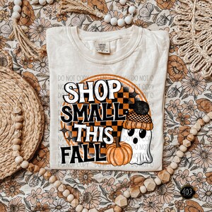 May include: A white t-shirt with a graphic design of a ghost wearing a black and white checkered hat and holding a pumpkin. The graphic also includes the text "Shop Small This Fall" in a black and orange circle.