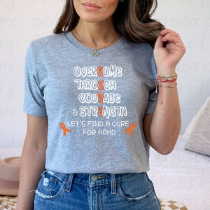 May include: A gray t-shirt with the text "Overcome Through Courage & Strength Let's Find A Cure For ADHD" in white. Two orange awareness ribbons are on either side of the text.