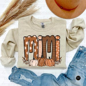 May include: A beige sweatshirt with the word "Mimi" in brown, pink, and white patterned letters. The letters are decorated with small pumpkins and leaves.