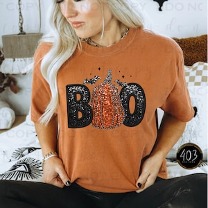 May include: A burnt orange t-shirt with a black sparkly "BOO" graphic featuring a pumpkin with bats. The pumpkin is orange with black glitter accents.