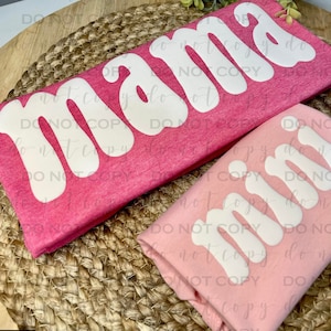 Op de afbeelding: Twee roze T-shirts met het woord "mama" in witte, gestileerde letters. De shirts zijn gevouwen en op elkaar gestapeld.