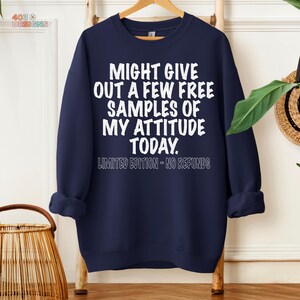 May include: Navy blue sweatshirt with white text that reads "MIGHT GIVE OUT A FEW FREE SAMPLES OF MY ATTITUDE TODAY." Below the text is "LIMITED EDITION - NO REFUNDS." The sweatshirt is hanging on a wooden rack.