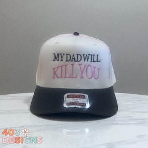 May include: A white and black baseball cap with the embroidered text "MY DAD WILL KILL YOU" in black and pink. The cap has a flat brim and a small, dark purple button on top. The brand label "OTTO" is visible.