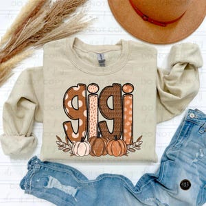 May include: A beige sweatshirt with the word "gigi" in brown, pink, and white patterned letters. The letters are decorated with polka dots and other patterns. Below the word "gigi" are three brown, pink, and orange pumpkins with green leaves.