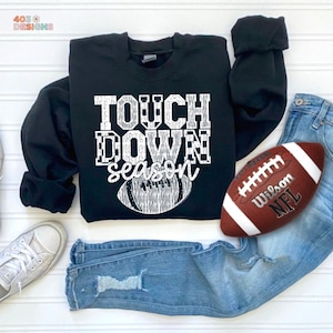 May include: Black sweatshirt with the text "TOUCH DOWN season" in white. A brown and white football is next to the sweatshirt, along with a pair of blue jeans and a pair of white sneakers. The sweatshirt is a crew neck.