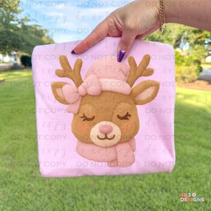 May include: A light pink tote bag featuring a felt applique of a reindeer with a pink hat, bow, and scarf. The reindeer has closed eyes and rosy cheeks. The bag is held up by a hand.