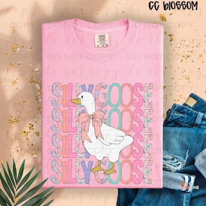 May include: A light pink Comfort Colors t-shirt featuring a cartoon goose with a pink bow and the text "SILLY GOOSE" in a colorful, layered design. The shirt is paired with blue jeans.