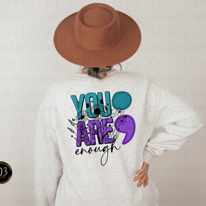 May include: A light gray sweatshirt with the words "YOU ARE enough" in teal, purple, and black. The design includes floral accents and a semicolon symbol. A brown hat is worn by the person wearing the sweatshirt.