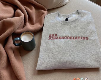 May include: A heather gray sweatshirt with the text "BRB. DISASSOCATING" in red. A dark teal mug with a white coffee drink is next to a folded brown blanket. The sweatshirt is folded on a beige surface.