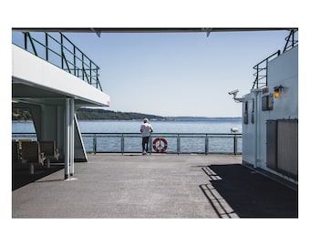 May include: A person stands on the deck of a ferry, looking out at the water. The ferry has white walls, green railings, and a red and white life preserver. The sky is blue, and the water is calm.
