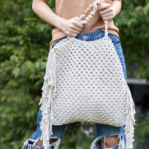 May include: A white crocheted tote bag with fringe detail and wooden bead accents. The bag is being held by a person wearing blue jeans.