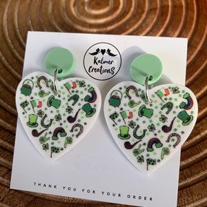May include: Heart-shaped earrings with a St. Patrick's Day theme. The white earrings feature green shamrocks, hats, horseshoes, and rainbows. The earrings have a light green circular top and silver hooks. The card behind the earrings says "Thank you for your order".