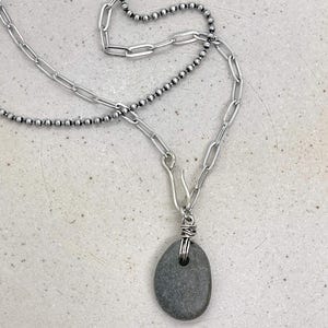 May include: A silver necklace featuring a gray, oval-shaped stone pendant. The necklace has two chain styles: a beaded chain and a link chain. The pendant is wrapped with silver wire and hangs from a hook clasp.
