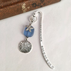 May include: Silver bookmark with a blue glass bead, a silver starfish charm, and a silver sand dollar charm. The bookmark has a decorative edge and a letter "C" charm.