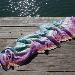 May include: A colorful crocheted shawl with a gradient of blues, greens, pinks, and yellows. The shawl is draped over a wooden dock with a rope and a body of water in the background.