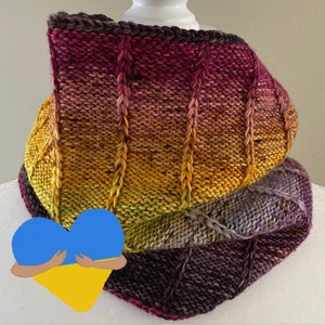 May include: A hand-knitted cowl in a gradient of colours, transitioning from yellow to purple. The cowl features vertical ribbed detailing and is displayed on a white mannequin head. The yarn has a speckled texture, adding visual interest to the design.