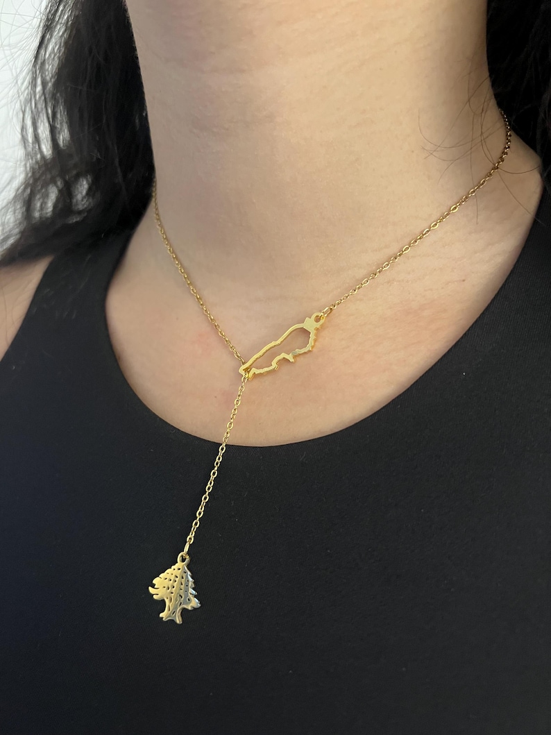 May include: A gold-toned lariat necklace featuring a delicate chain. The necklace includes a cutout pendant shaped like the map of Lebanon and a dangling charm in the shape of a cedar tree, both suspended from a thin, polished gold-coloured metal chain.