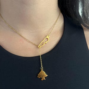 May include: A gold-toned necklace featuring a delicate chain with a cutout pendant shaped like the map of Lebanon, connected to a vertical drop chain that ends in a small, detailed cedar tree charm with a polished metallic finish.