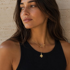 May include: A gold-toned lariat necklace featuring a delicate chain, a small outline of the country of Lebanon, and a dangling charm shaped like a cedar tree. The necklace rests against the neck, with the chain extending downward to the tree charm.
